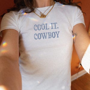 May include: White t-shirt with the text "COOL IT. COWBOY" in light blue. The shirt is a crew neck style. The model is wearing a gold necklace and blue jeans. The background is a warm, orange-toned wall.