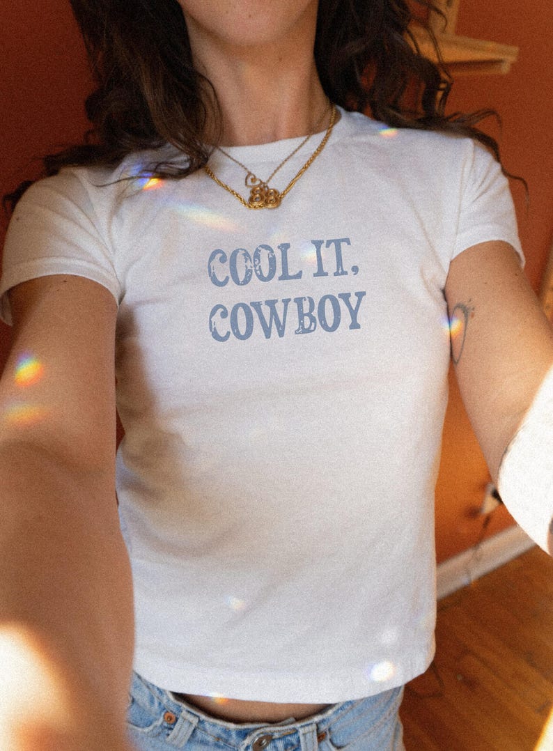 May include: White t-shirt with the text "COOL IT. COWBOY" in light blue. The shirt is a crew neck style. The model is wearing a gold necklace and blue jeans. The background is a warm, orange-toned wall.