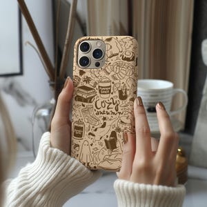 May include: A beige phone case with a repeating autumn-themed pattern. The design includes illustrations of a pumpkin spice latte, books, a cosy sock, and the words "Cozy Autumn". The case is decorated with leaves and other seasonal elements.