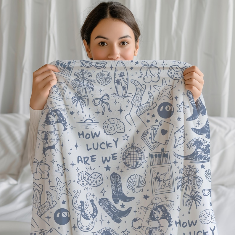 May include: A white throw blanket with a blue and white pattern. The pattern includes palm trees, stars, cherries, dice, horseshoes, a disco ball, a surfboard, and the text "How lucky are we".