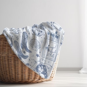 May include: A white throw blanket with a blue and white pattern of cowboy boots, dice, a disco ball, a seashell, matches, and the text "Cowgirl Beach Club". The blanket is folded and placed in a wicker basket.