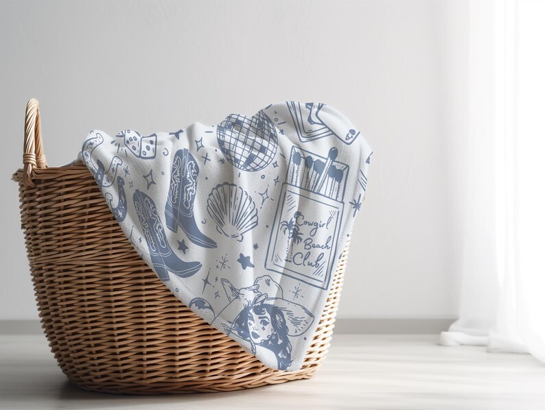 May include: A white throw blanket with a blue and white pattern of cowboy boots, dice, a disco ball, a seashell, matches, and the text "Cowgirl Beach Club". The blanket is folded and placed in a wicker basket.