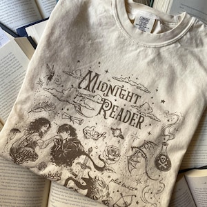 Puede incluir: Una camiseta color crema con ilustraciones en tinta marr&oacute;n oscuro y el texto "Midnight Reader" y "plot twist". El gr&aacute;fico incluye una pareja, una espada, una rosa, una botella con una calavera, un drag&oacute;n, una m&aacute;scara y una pluma.
