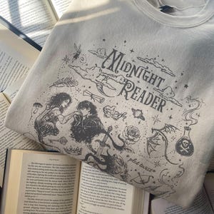 Op de afbeelding: Een beige sweater met ronde hals en een zwarte grafische print met de tekst "Midnight Reader" en "plot twist". Het ontwerp bevat illustraties van een koppel, een zwaard, een roos, een toverdrankflesje, een drakenvleugel, wolken en sterren.