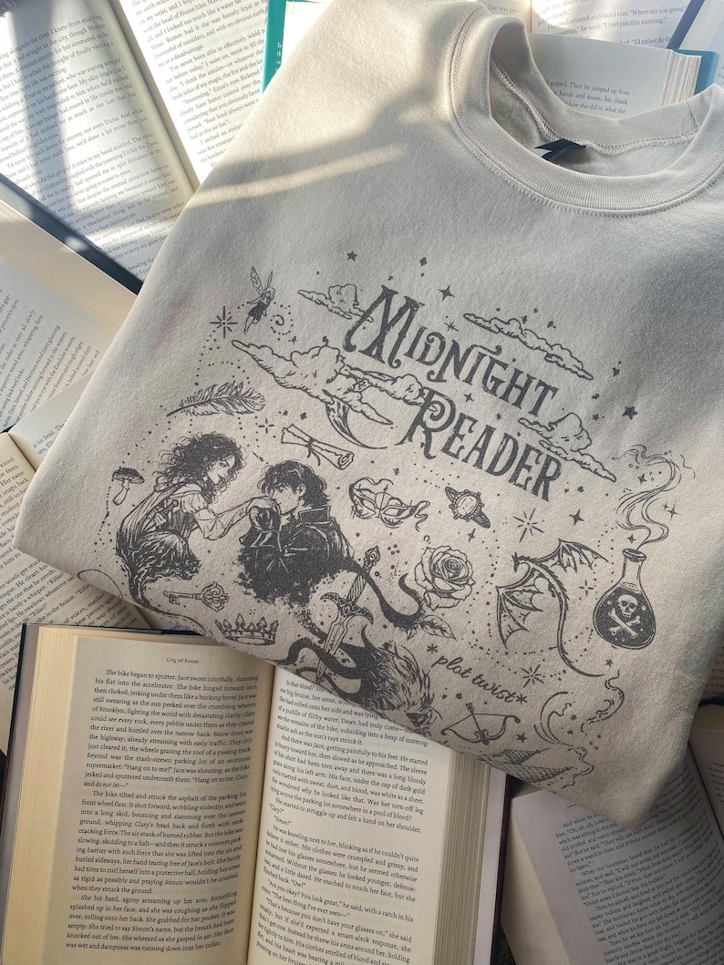 Op de afbeelding: Een beige sweater met ronde hals en een zwarte grafische print met de tekst "Midnight Reader" en "plot twist". Het ontwerp bevat illustraties van een koppel, een zwaard, een roos, een toverdrankflesje, een drakenvleugel, wolken en sterren.