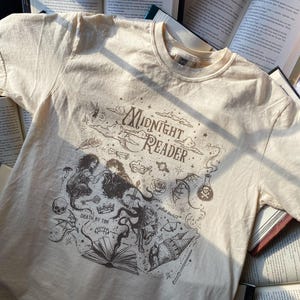 Puede incluir: Una camiseta de manga corta color crema con un estampado gr&aacute;fico marr&oacute;n oscuro con el texto Midnight Reader, Death by TBR y plot twist. El dise&ntilde;o incluye ilustraciones de un libro, una calavera, una poci&oacute;n, una corona y un drag&oacute;n.