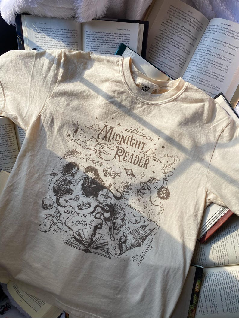 Puede incluir: Una camiseta de manga corta color crema con un estampado gr&aacute;fico marr&oacute;n oscuro con el texto Midnight Reader, Death by TBR y plot twist. El dise&ntilde;o incluye ilustraciones de un libro, una calavera, una poci&oacute;n, una corona y un drag&oacute;n.
