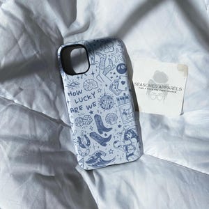 Puede incluir: Funda de tel&eacute;fono azul claro con un patr&oacute;n repetido de ilustraciones en azul oscuro, que incluyen botas, dados y una bola de discoteca. Tambi&eacute;n aparece el texto "HOW LUCKY ARE WE". Una peque&ntilde;a tarjeta con el texto "SEASONED APPARELS" est&aacute; al lado.