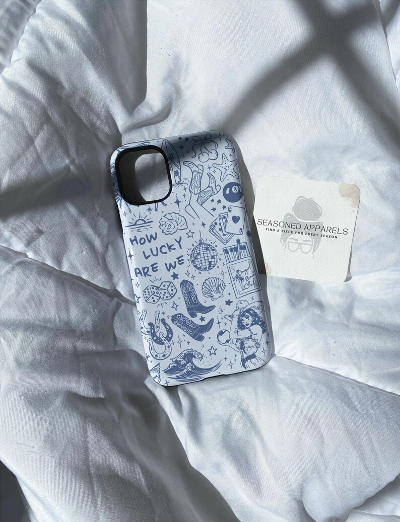 Puede incluir: Funda de tel&eacute;fono azul claro con un patr&oacute;n repetido de ilustraciones en azul oscuro, que incluyen botas, dados y una bola de discoteca. Tambi&eacute;n aparece el texto "HOW LUCKY ARE WE". Una peque&ntilde;a tarjeta con el texto "SEASONED APPARELS" est&aacute; al lado.