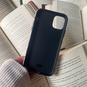 May include: A dark blue phone case with the text "IP11" is held over open books. The case has a smooth, matte finish and is designed to fit a specific phone model. The background features pages of a book.