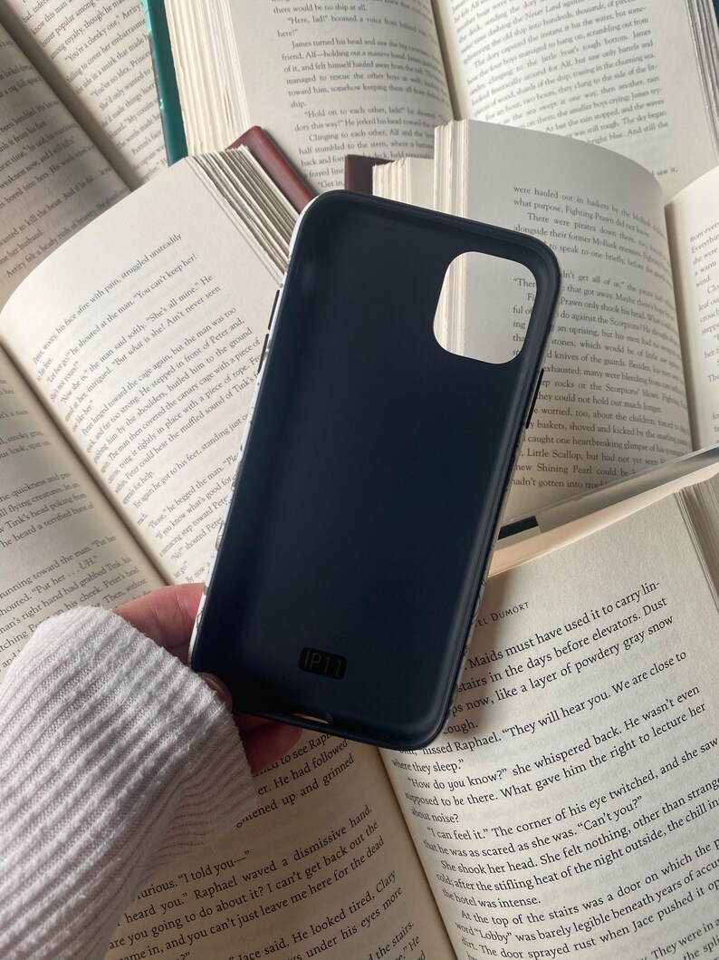 May include: A dark blue phone case with the text "IP11" is held over open books. The case has a smooth, matte finish and is designed to fit a specific phone model. The background features pages of a book.