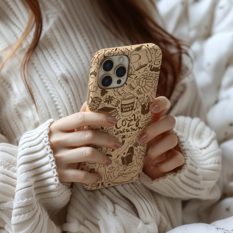 May include: A beige phone case with a brown doodle design featuring autumn themes. The case includes drawings of a coffee cup, a book, and the words "Pumpkin Spice" and "Cozy Autumn". The case is designed for a smartphone.