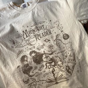 Puede incluir: Una camiseta de manga corta color crema con un estampado gr&aacute;fico marr&oacute;n oscuro que incluye el texto "MIDNIGHT READER", "*plot twist*" y "DEATH BY TBR". El dise&ntilde;o presenta ilustraciones de una pareja, un lobo, un castillo, una calavera y una poci&oacute;n.