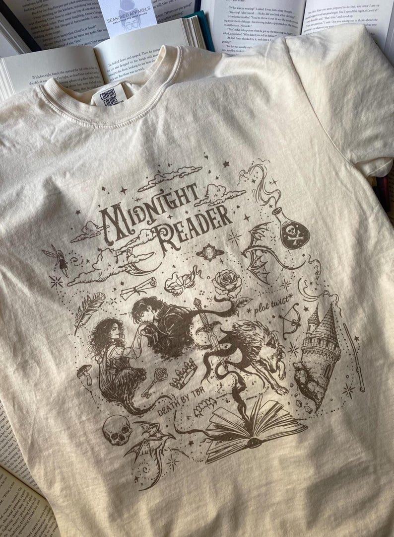 Puede incluir: Una camiseta de manga corta color crema con un estampado gr&aacute;fico marr&oacute;n oscuro que incluye el texto "MIDNIGHT READER", "*plot twist*" y "DEATH BY TBR". El dise&ntilde;o presenta ilustraciones de una pareja, un lobo, un castillo, una calavera y una poci&oacute;n.