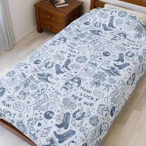 May include: A white twin-sized bedspread with a blue and white pattern of horseshoes, dice, stars, and palm trees. The pattern repeats the phrase "How lucky are we?" throughout the bedspread.