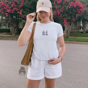 May include: A person wearing a light blue t-shirt with the number "21" in black, paired with white shorts and a beige baseball cap. The person is also wearing gold earrings and a brown shoulder bag. The background features pink flowering trees.