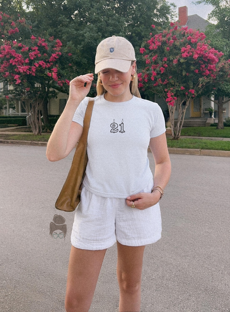 May include: A person wearing a light blue t-shirt with the number "21" in black, paired with white shorts and a beige baseball cap. The person is also wearing gold earrings and a brown shoulder bag. The background features pink flowering trees.