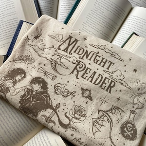 Puede incluir: Un art&iacute;culo de tela beige con ilustraciones serigrafiadas en marr&oacute;n oscuro, incluyendo el texto "MIDNIGHT READER" y "plot twist". El dise&ntilde;o incluye una pareja, una m&aacute;scara, una rosa, un drag&oacute;n, una botella con una calavera, estrellas y nubes.