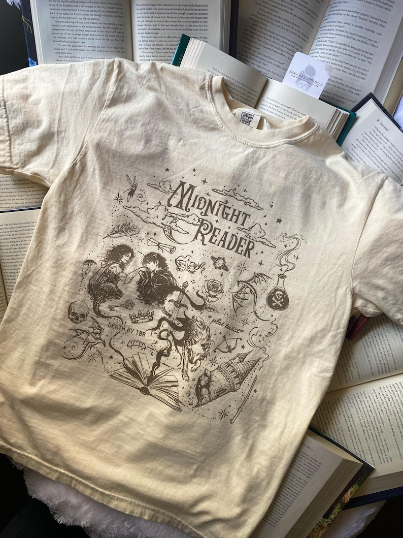 Puede incluir: Una camiseta de manga corta color crema con un estampado gr&aacute;fico marr&oacute;n oscuro con el texto Midnight Reader, Death by TBR y plot twist. El dise&ntilde;o incluye ilustraciones de una calavera, una rosa, un frasco de poci&oacute;n, un drag&oacute;n y un castillo.