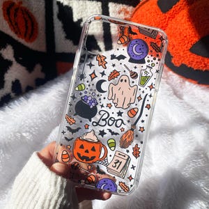 Könnte beinhalten: Transparente Handyhülle mit Halloween-Design. Die Hülle zeigt Illustrationen eines Geistes, eines Kürbisbechers, eines Hexenbesens und das Wort "Boo". Weitere Elemente sind Fledermäuse, Süßigkeiten und ein Kalenderblatt.
