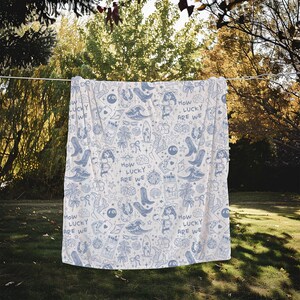 May include: A white throw blanket with a blue and white pattern of various symbols, including horseshoes, palm trees, and the text "How Lucky Are We".