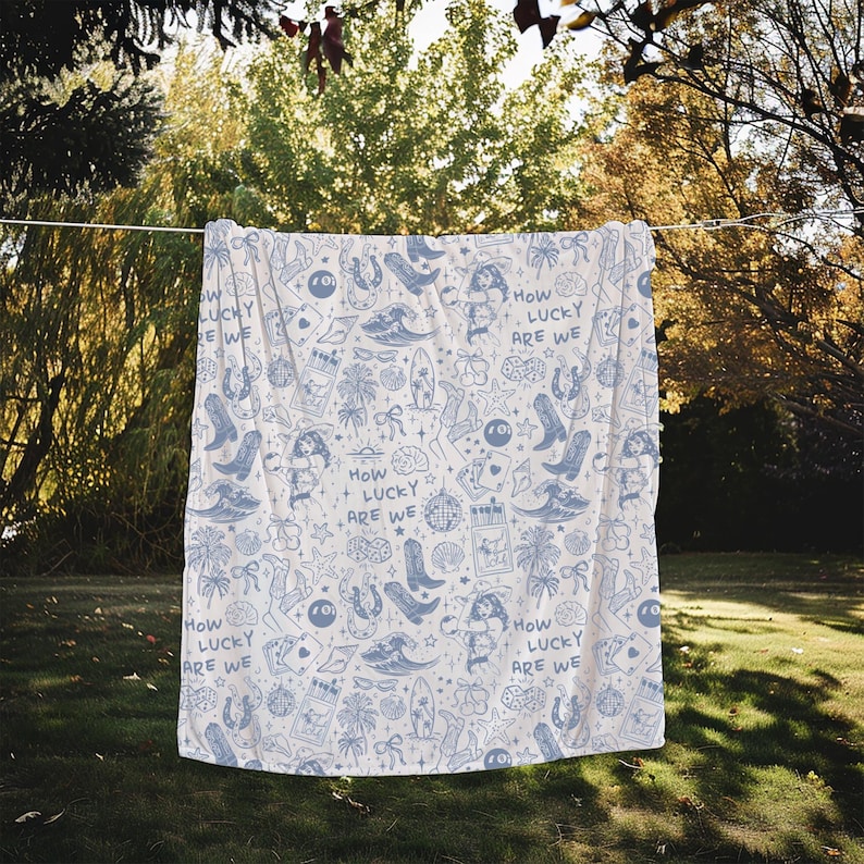 May include: A white throw blanket with a blue and white pattern of various symbols, including horseshoes, palm trees, and the text "How Lucky Are We".