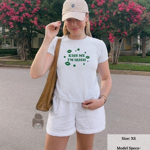 May include: A light gray t-shirt with the text "KISS ME I'M IRISH" in green, paired with white shorts and a beige baseball cap. The outfit is completed with a brown shoulder bag. The image also includes text that says "Size: XS".