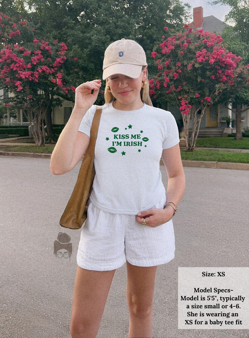 May include: A light gray t-shirt with the text "KISS ME I'M IRISH" in green, paired with white shorts and a beige baseball cap. The outfit is completed with a brown shoulder bag. The image also includes text that says "Size: XS".