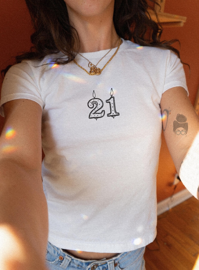 May include: White t-shirt with a black and white illustration of the number "21" in a candle design. The candles are lit with flames. The shirt is short-sleeved and has a round neckline.