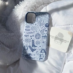 Puede incluir: Funda de tel&eacute;fono azul claro con un dise&ntilde;o de garabatos blanco con botas de vaquero, dados, una bola de discoteca y una mujer. El texto "HOW LUCKY ARE WE" tambi&eacute;n est&aacute; en la funda. Una posavasos con el texto "SEASONED APPARELS" est&aacute; al lado.