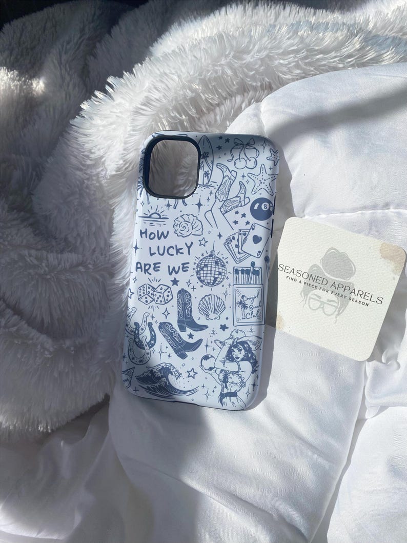 Puede incluir: Funda de tel&eacute;fono azul claro con un dise&ntilde;o de garabatos blanco con botas de vaquero, dados, una bola de discoteca y una mujer. El texto "HOW LUCKY ARE WE" tambi&eacute;n est&aacute; en la funda. Una posavasos con el texto "SEASONED APPARELS" est&aacute; al lado.