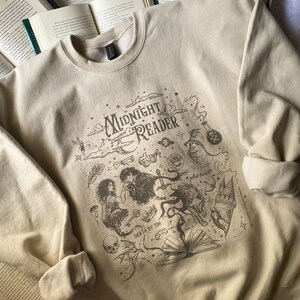 Op de afbeelding: Een beige sweater met ronde hals en een donkerbruine grafische print met de tekst "MIDNIGHT READER" en "DEATH BY TBR". Het ontwerp bevat illustraties van een persoon, een wolf, een kasteel, een schedel, een roos en diverse sterren.