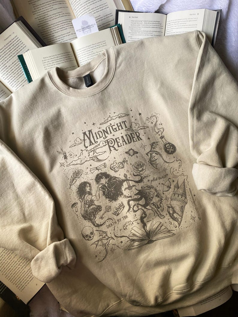 Op de afbeelding: Een beige sweater met ronde hals en een donkerbruine grafische print met de tekst "MIDNIGHT READER" en "DEATH BY TBR". Het ontwerp bevat illustraties van een persoon, een wolf, een kasteel, een schedel, een roos en diverse sterren.