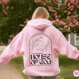 Puede incluir: Una sudadera con capucha rosa claro con un estampado gr&aacute;fico negro en la espalda. El gr&aacute;fico presenta un olivo con el texto "I am like an olive tree flourishing in the HOUSE OF GOD".