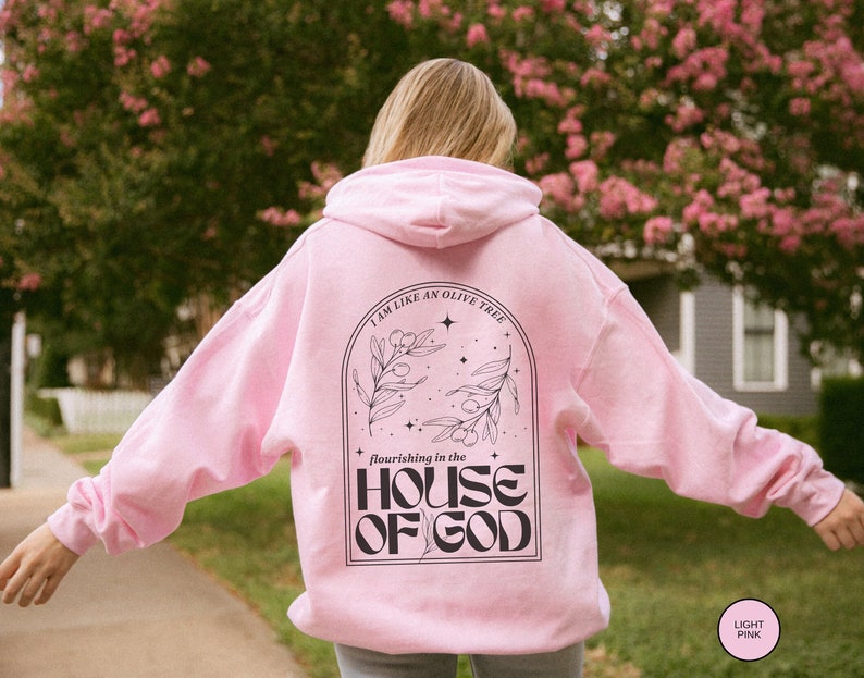 Puede incluir: Una sudadera con capucha rosa claro con un estampado gr&aacute;fico negro en la espalda. El gr&aacute;fico presenta un olivo con el texto "I am like an olive tree flourishing in the HOUSE OF GOD".