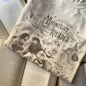 Puede incluir: Una camiseta color crema con un estampado gr&aacute;fico marr&oacute;n oscuro con el texto Midnight Reader y plot twist. El dise&ntilde;o incluye ilustraciones de una pareja, una espada, una rosa, una poci&oacute;n, un hada, una m&aacute;scara y estrellas en la tela.