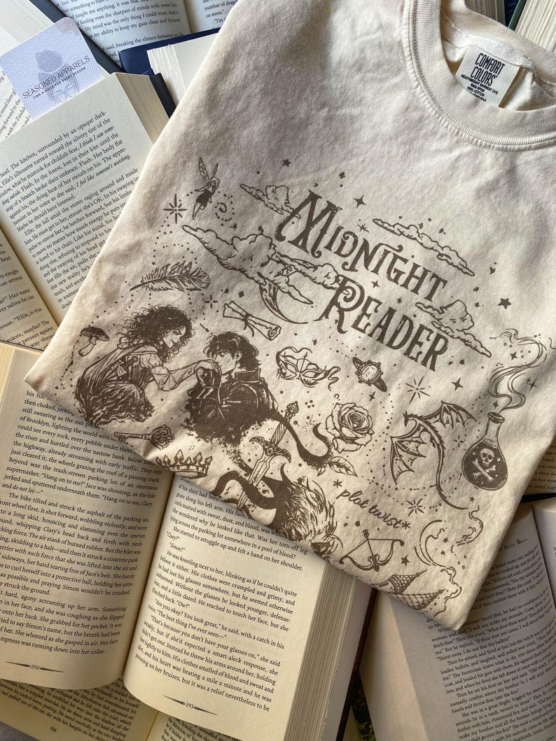 Puede incluir: Una camiseta color crema con un estampado gr&aacute;fico marr&oacute;n oscuro con el texto Midnight Reader y plot twist. El dise&ntilde;o incluye ilustraciones de una pareja, una espada, una rosa, una poci&oacute;n, un hada, una m&aacute;scara y estrellas en la tela.