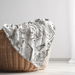 May include: A white blanket with a black and white print of books, stars, and a ticket that says "Welcome to the Fantasy Book Club, Issue One". The blanket is folded and placed in a wicker basket.