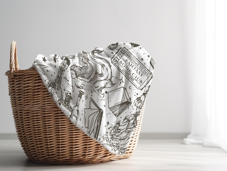 May include: A white blanket with a black and white print of books, stars, and a ticket that says "Welcome to the Fantasy Book Club, Issue One". The blanket is folded and placed in a wicker basket.