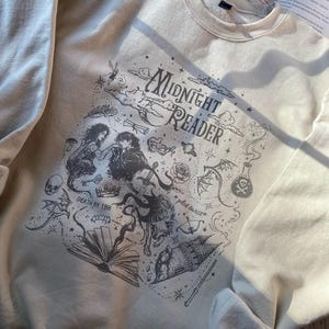 Op de afbeelding: Een cr&egrave;mekleurige sweater met ronde hals en een donkergrijze grafische print met de tekst "Midnight Reader", "Death by TBR" en "plot twist". Het ontwerp bevat illustraties van een koppel, een boek, een kasteel, een draak en een schedel.