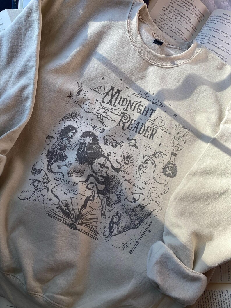 Op de afbeelding: Een cr&egrave;mekleurige sweater met ronde hals en een donkergrijze grafische print met de tekst "Midnight Reader", "Death by TBR" en "plot twist". Het ontwerp bevat illustraties van een koppel, een boek, een kasteel, een draak en een schedel.
