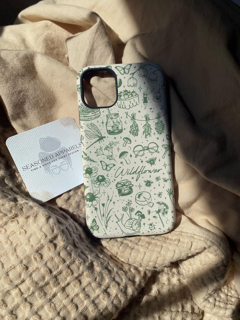 Może przedstawiać: Etui na telefon z kremowym tłem i zielonym wzorem polnych kwiat&oacute;w. Wz&oacute;r zawiera motyle, kwiaty, słoiki i słowo "Wildflower". Widoczna jest r&oacute;wnież wizyt&oacute;wka z napisem "SEASONED APPARELS".