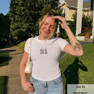 May include: White t-shirt with the number "21" printed in black. The model is wearing an XS size. The model is 5'5" and typically wears a size small or 4-6. She is wearing an XS for a baby tee fit.
