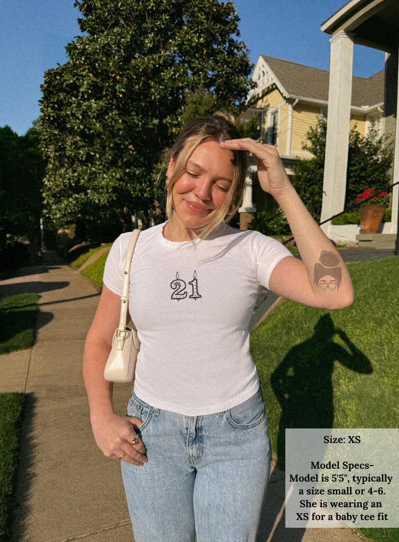 May include: White t-shirt with the number "21" printed in black. The model is wearing an XS size. The model is 5'5" and typically wears a size small or 4-6. She is wearing an XS for a baby tee fit.