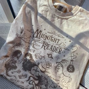 Puede incluir: Una camiseta de cuello redondo color crema con ilustraciones gr&aacute;ficas en marr&oacute;n oscuro que incluyen el texto "Midnight Reader", "Death by TBR" y "plot twist". El dise&ntilde;o muestra una pareja, una espada, una poci&oacute;n, una rosa y una corona.