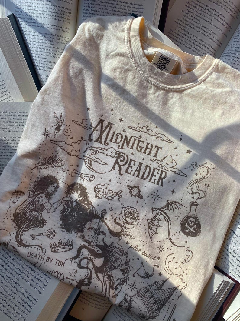 Puede incluir: Una camiseta de cuello redondo color crema con ilustraciones gr&aacute;ficas en marr&oacute;n oscuro que incluyen el texto "Midnight Reader", "Death by TBR" y "plot twist". El dise&ntilde;o muestra una pareja, una espada, una poci&oacute;n, una rosa y una corona.