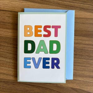 May include: A greeting card with the words "BEST DAD EVER" in colorful block letters. The card is white with a light green border and is paired with a light blue envelope. The card and envelope are on a wooden surface.