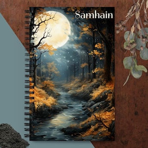 Puede incluir: Un cuaderno de espiral con una imagen a todo color de una escena de bosque iluminado por la luna. La imagen presenta un arroyo sinuoso, árboles con hojas doradas y una gran luna llena en el cielo. El texto "Samhain" está impreso en blanco en la parte superior del cuaderno.