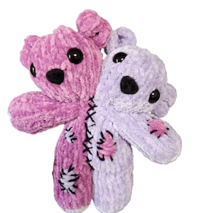 May include: A handcrafted plush toy depicting two teddy bears joined at the seam. One is a bright pink, the other a soft lavender. Both have black button eyes and stitched details, with patches on their limbs.
