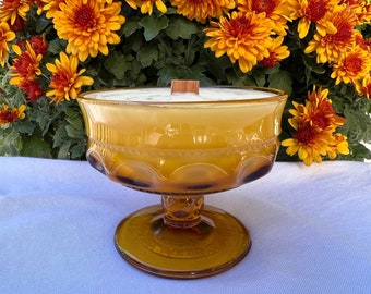 May include: An amber glass candle holder with a white candle and a wooden wick. The candle holder has a pedestal base and a decorative pattern.